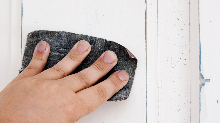 a hand sanding the surface of a door with a piece of sand paper