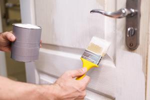 A person holding a tin of paint and a brush, whilst finishing a door